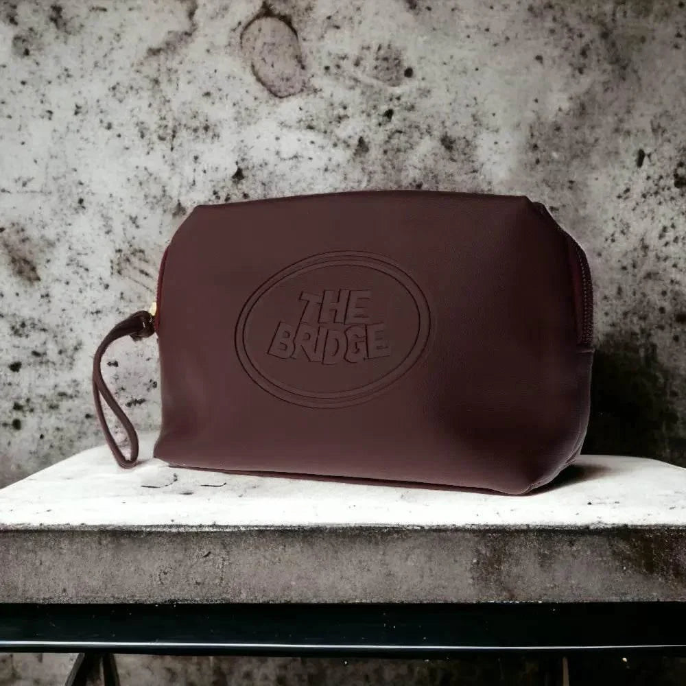 Braune Kulturtasche aus Leder mit geprägtem 'The Bridge' Logo auf Steinoberfläche