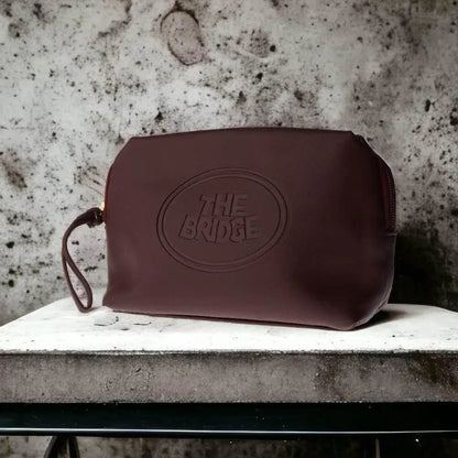 Braune Kulturtasche aus Leder mit geprägtem 'The Bridge' Logo auf Steinoberfläche