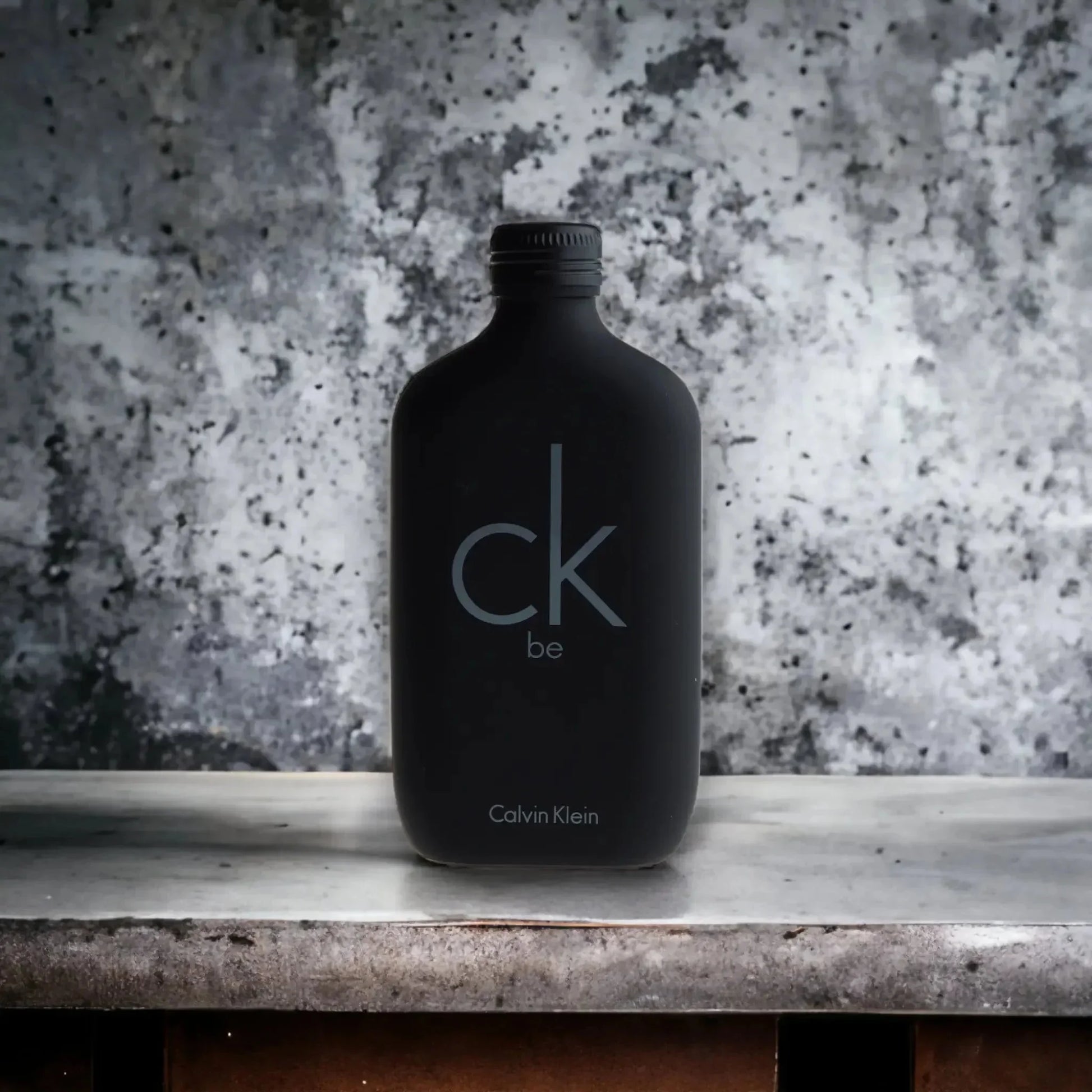 Schwarzer Flakon ck be Eau de Toilette steht auf Marmoroberfläche vor grauem Hintergrund.