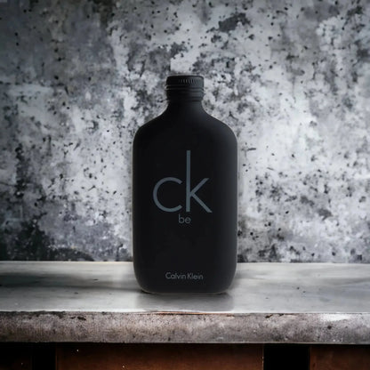 Schwarzer Flakon ck be Eau de Toilette steht auf Marmoroberfläche vor grauem Hintergrund.