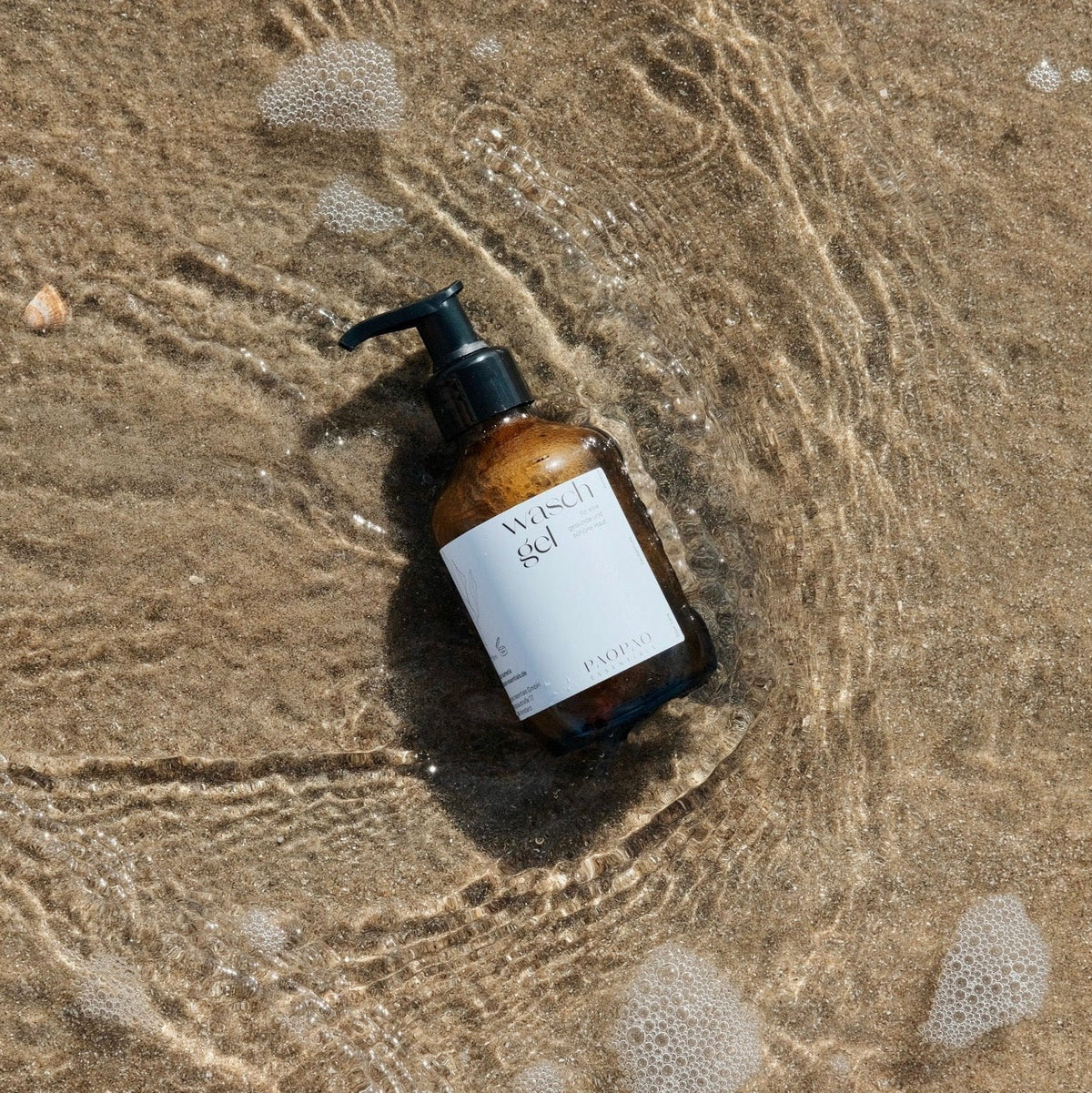 paopao essentials Waschgel in einer braunen Flasche mit Pumpspender im flachen Wasser am Strand.