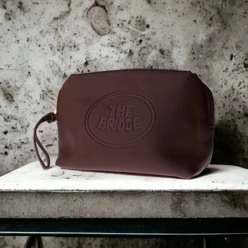 Braune Leder-Kosmetiktasche auf Steinuntergrund, geprägt mit 'The Bridge'-Logo, schlichtes Design.